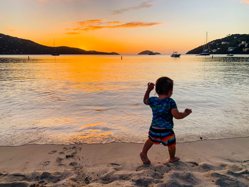 A toddler is walking on the beach at sunset.