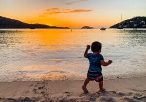 A toddler is walking on the beach at sunset.